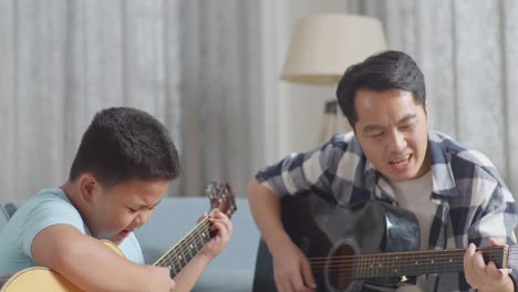 close up of asian father and son with expressive face enjoys singing and playing the guitar together on sofa at home