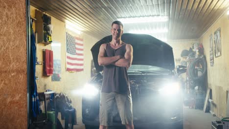 Portrait-of-a-man-with-a-short-haircut-in-a-T-shirt,-a-motorist-mechanic-who-stands-against-the-backdrop-of-his-workshop-garage-and-a-car-with-an-open-hood-and-burning-headlights