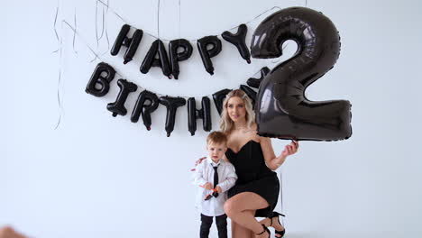 lady wearing a black dress kneeled near the little baby boy wearing white shirt, black tie and jeans. mother is holding a large balloon of number two shape. birthday footage in studio.