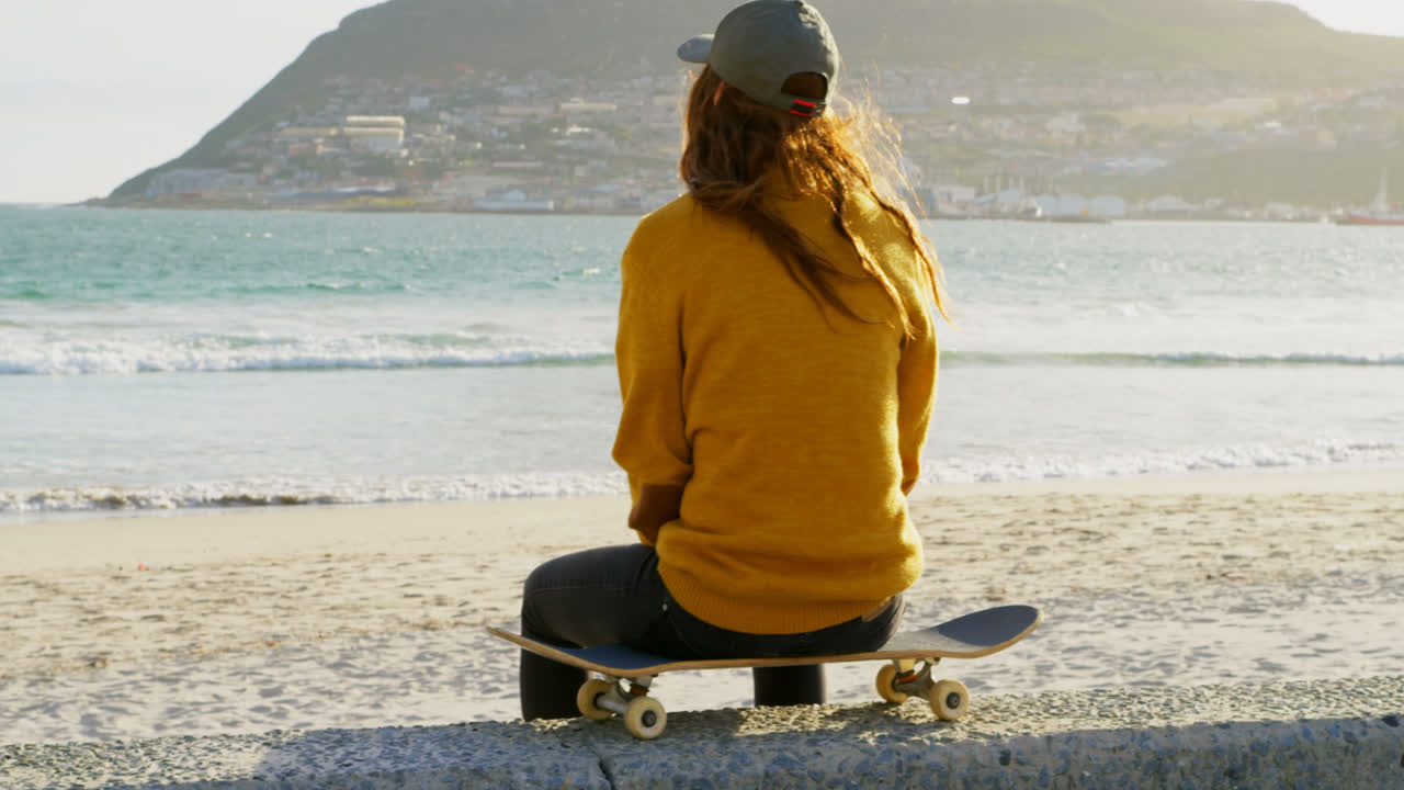 vista trasera de una joven mujer caucásica sentada en una patineta y mirando al mar en la playa 4k