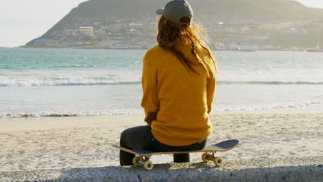 Rückansicht-Einer-Jungen-Kaukasischen-Frau,-Die-Auf-Einem-Skateboard-Sitzt-Und-Am-Strand-Auf-Das-Meer-Blickt,-4k