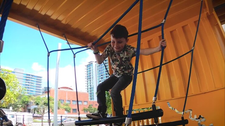 A happy young boy reaches out and climbs across a rope swing in an industrial playground