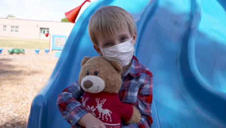 little boy in a mask cuddling his teddy bear at a playground