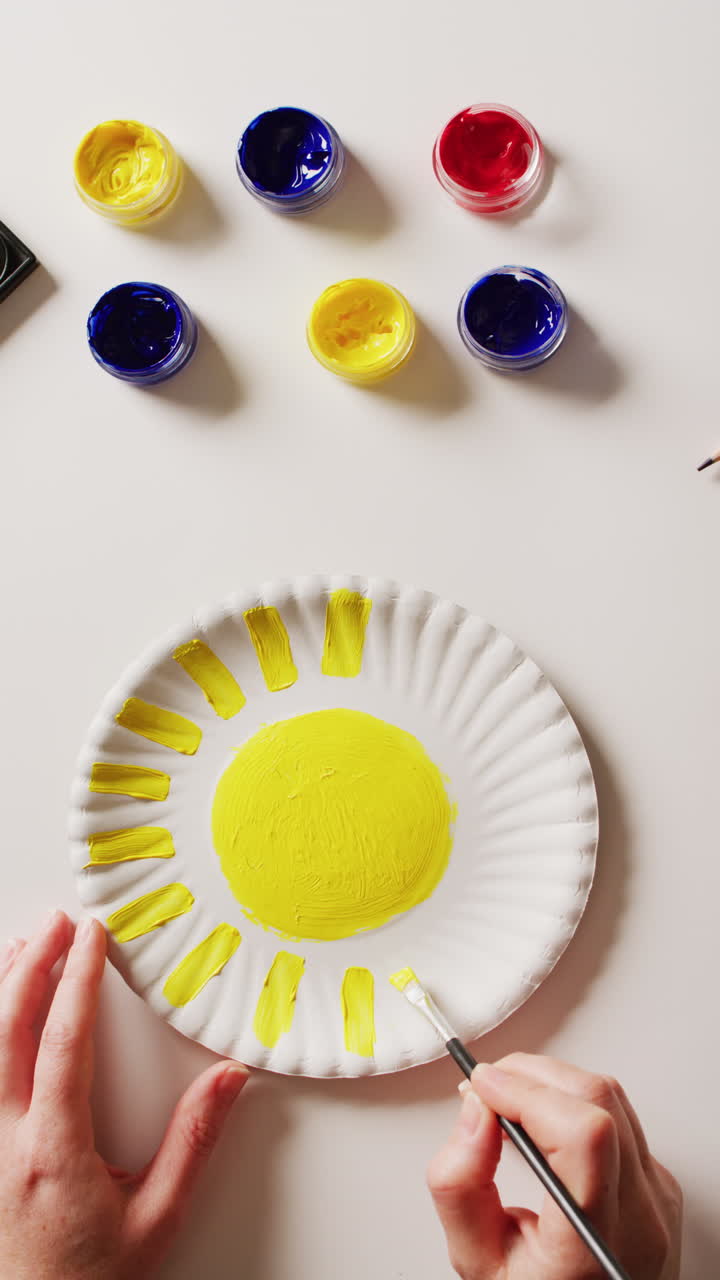 video vertical de manos pintando pintura amarilla en una placa de papel, con pinturas sobre un fondo blanco