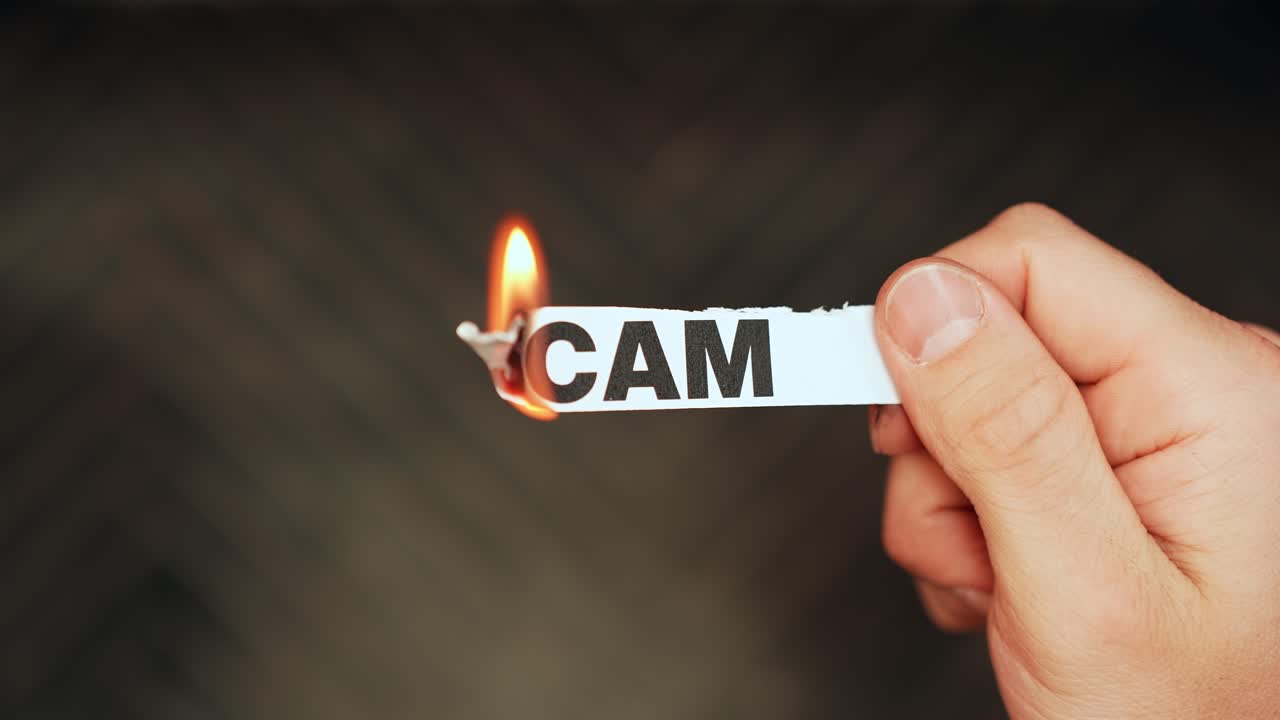 Primo piano di una mano che tiene una striscia di carta etichettata “SCAM” mentre il bordo brucia con la fiamma di un accendino su sfondo scuro, concetto di frode, inganno e rischio.