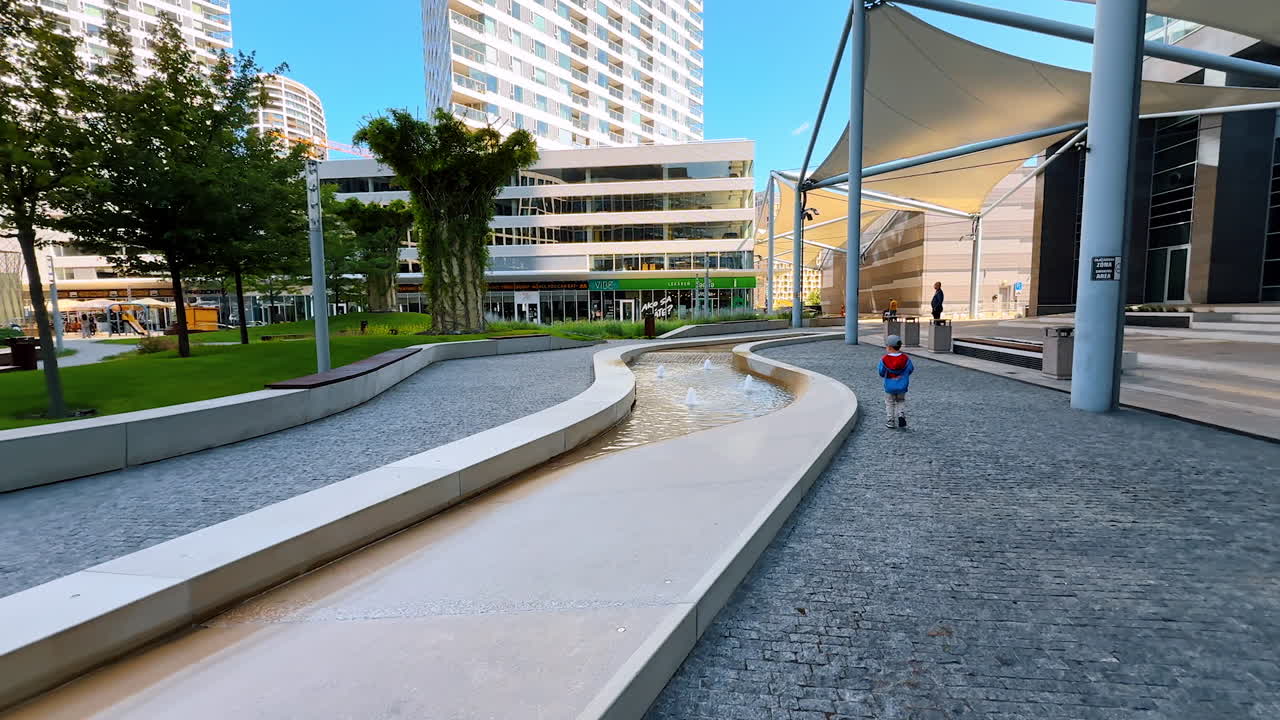 Kid runs on a plaza path. A child happily runs on a smooth path in a lively urban plaza filled with greenery and modern buildings