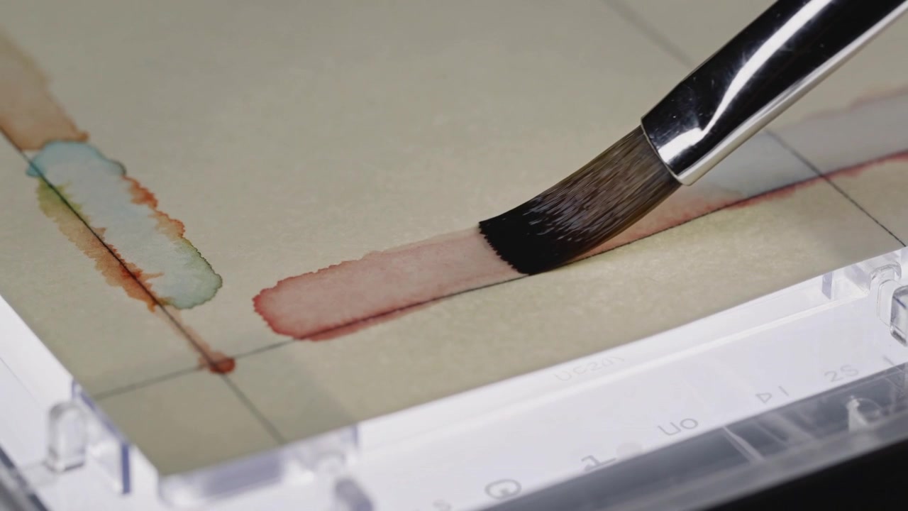 Dabbing artist brush pulling pink wash along grid paper, evening pigment with teal blot at desk