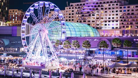 Close-Time-lapse-of-crowds-walking-around-fun-fare-while-Ferris-wheel-spins,-Darling-Harbour-Sydney,-Australia