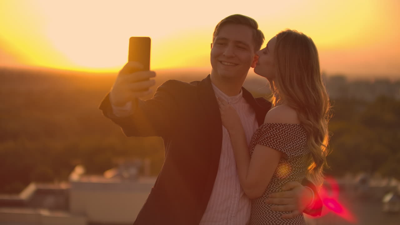 de pie en el techo al atardecer una pareja casada un hombre y una mujer se abrazan y se toman una selfie en el teléfono. toma fotos de ti mismo de pie en el tejado y abrazarse. jóvenes enamorados