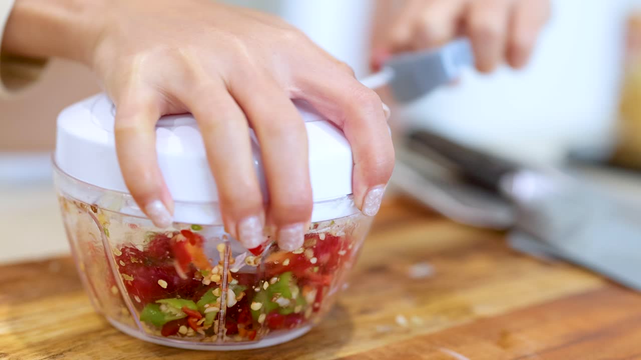 A hand operates a manual food processor, chopping vegetables on a wooden surface in a well-lit kitchen