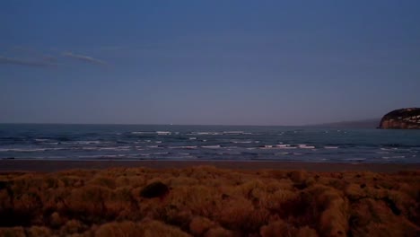 Antena-Baja-Sobre-Matorrales-Hacia-Una-Playa-Con-Olas-Al-Atardecer