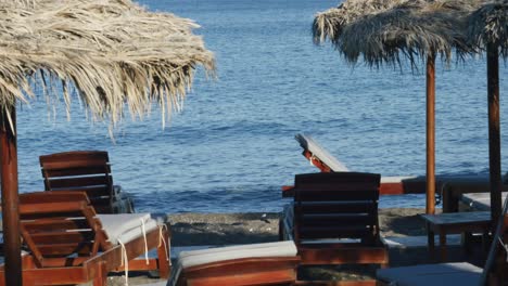close view of multiple relaxing lounge chairs on volcanic black sand at beach club near ocean in santorini, greece