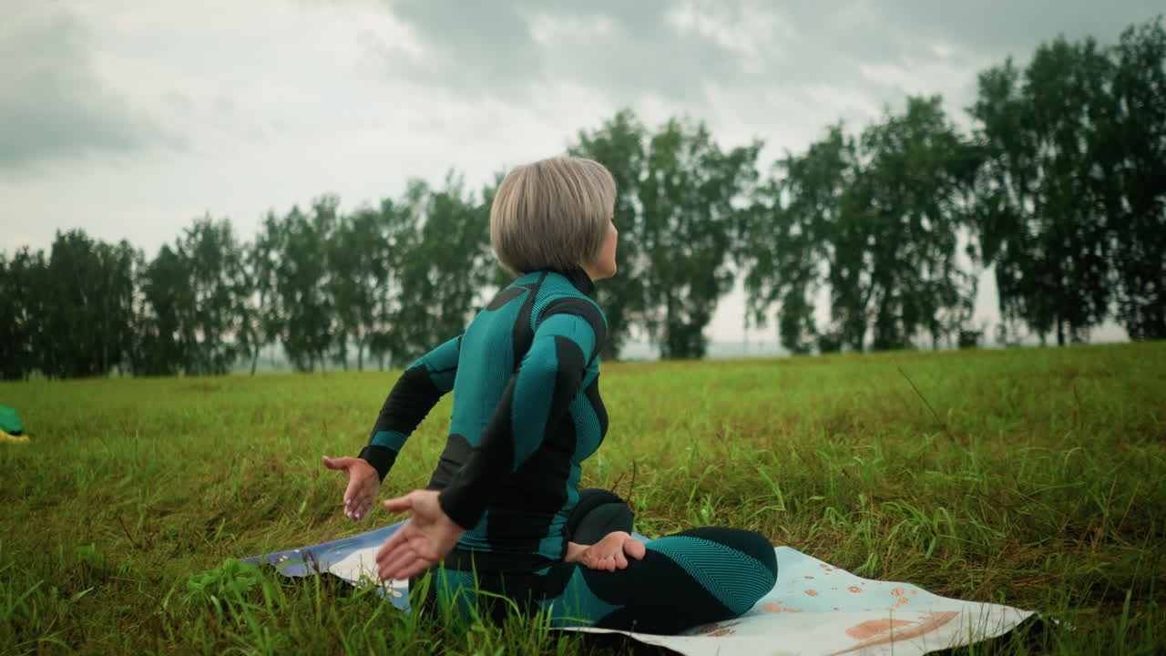 수카사나 포즈에서 매트에 앉아 눈을 감고 요가를 연습하는 중년 여성, 구름이 가득한 넓은 잔디에서, 배경에는 나무가 줄지어 있으며, 노란색 부츠와 옷이 근처에 있습니다.