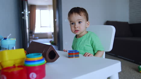 cute dark-haired boy sitting at the little desk. baby looks at a range of toys lying on the table.
