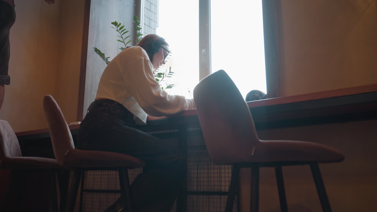 writer wearing headphones sits by window jotting notes in notepad with indoor plant and decorative berries beneath wall art in cozy ambient space