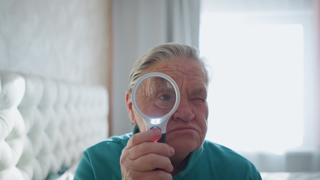 Elderly woman in green jacket holding magnifying glass, focusing on something through lens with serious expression, bright sunlight coming from window, relaxed indoor setting, seated on bed