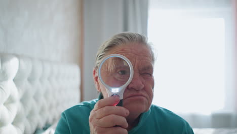 elderly woman in green jacket holding magnifying glass, focusing on something through lens with serious expression, bright sunlight coming from window, relaxed indoor setting, seated on bed