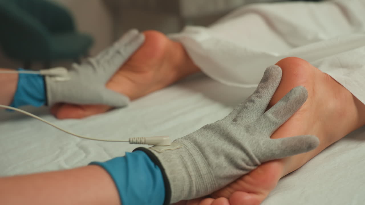 Mani guantate applicano microcorrente al piede, primo piano sul contatto degli elettrodi e sui fili, impulsi delicati e pressione tattile, ambiente di trattamento sterile, operatore concentrato che monitora la sensazione.