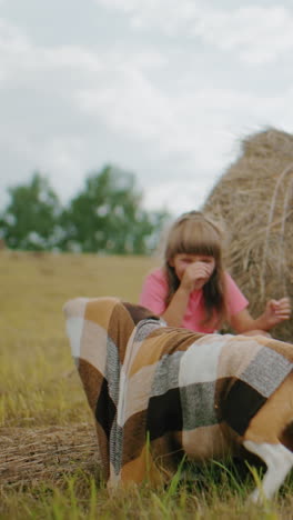 momento alegre como perro juguetón está cubierto con una manta, el dueño se ríe mientras mira, la hija se agacha cerca disfrutando de la interacción divertida, entorno al aire libre cálido con balas de heno y campo natural