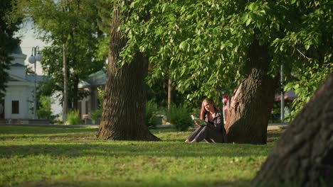 mujer sentada al aire libre bajo un árbol en un campo cubierto de hierba, leyendo un libro a la luz del sol cálida, las hojas de los árboles se balancean suavemente en la brisa, con el fondo mostrando un edificio residencial y un poste de lámpara