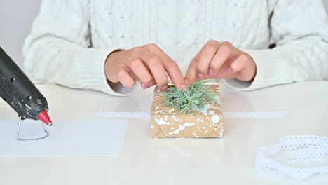 female hands uses a glue gun to decorate a wrapped gift with a pine cone and a branch