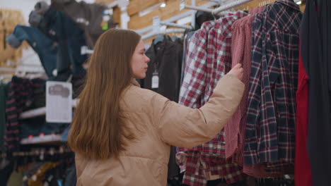 se ve a una chica con una chaqueta marrón y pantalones negros examinando de cerca y tocando la ropa colgada en un estante en una tienda. un momento de compras mientras navega a través de una selección de camisas a cuadros