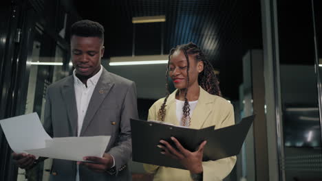 Two-business-partners-a-black-man-and-a-woman-walk-down-the-hallway-of-the-office-and-discuss-papers.-A-business-meeting-on-the-go.-Discuss-reports-while-walking-in-the-office