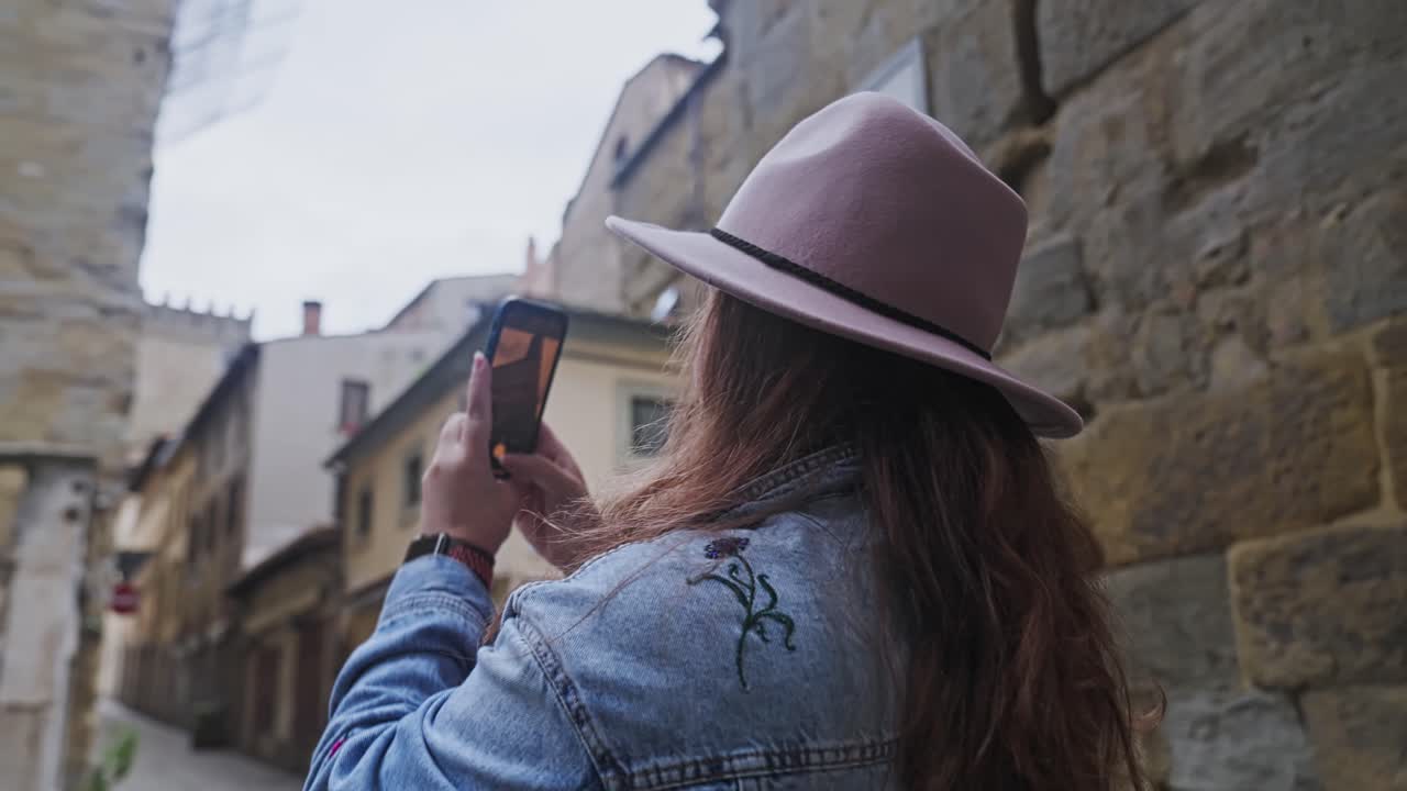 hermosa mujer turista con chaqueta de mezclilla y sombrero tomando fotos de la arquitectura italiana histórica con la cámara de su teléfono inteligente