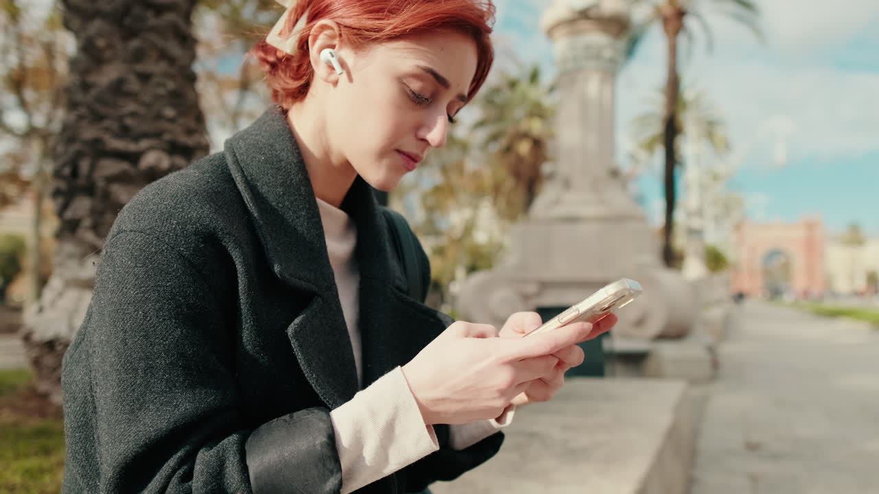 Young Redhead Woman Using Smartphone with Earbuds in Park
