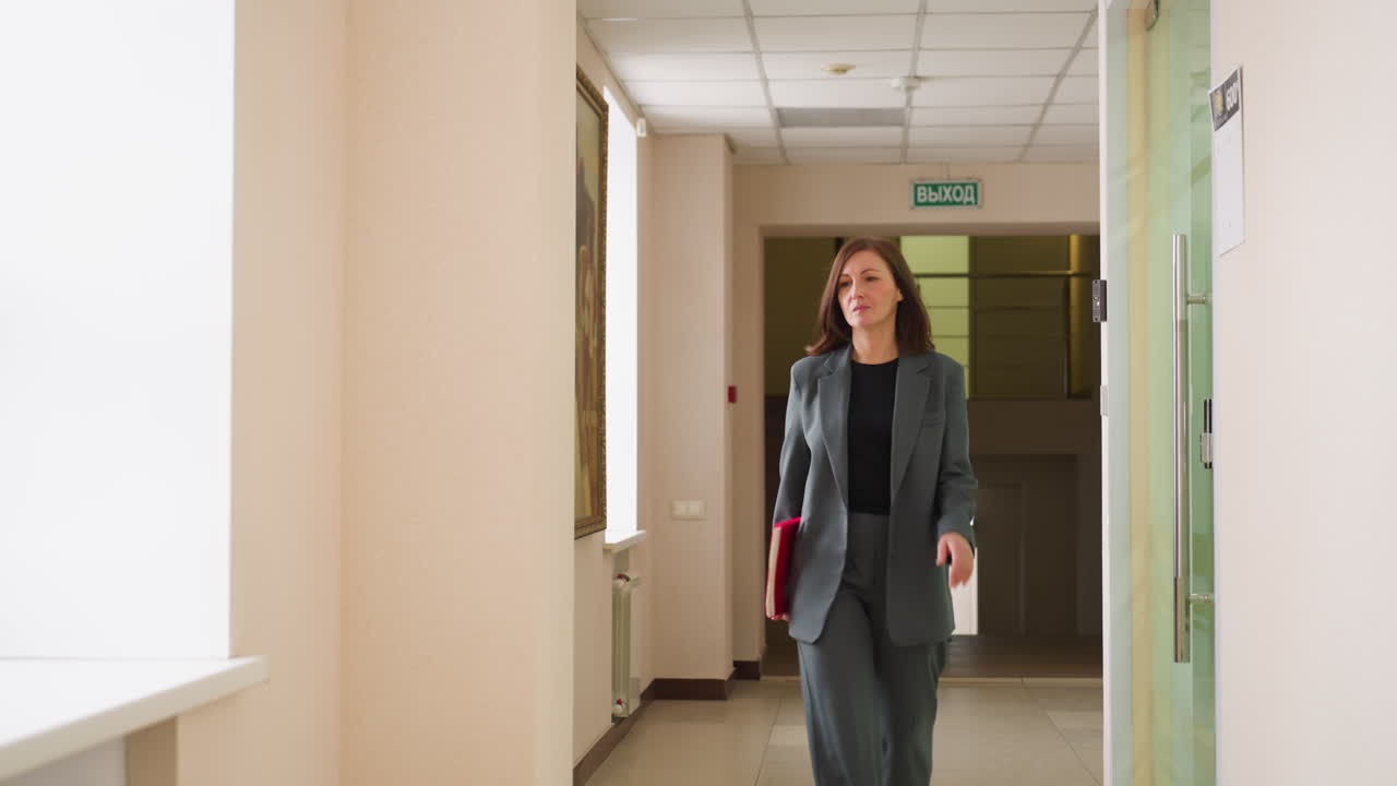 Confident businesswoman in gray suit walking through bright office corridor holding red folder and coffee cup, heading toward camera with determined expression in corporate workplace environment