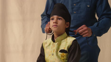 Studio-Shot-Of-Father-Tying-Turban-Onto-Head-Of-Young-Sikh-Son-Wearing-Traditional-Waistcoat-As-Sequence-Part-2-Of-10