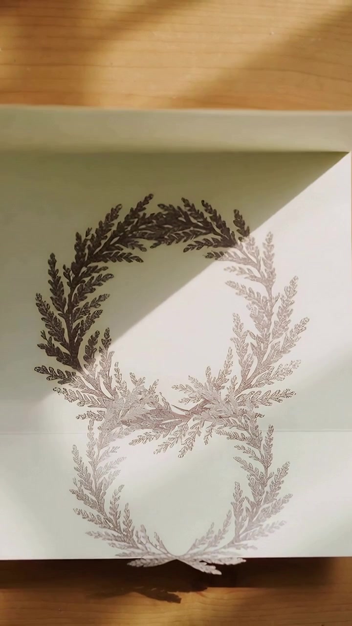 Vertical video: Lifting top flap of folded cream card revealing inner laurel wreaths on wood desk