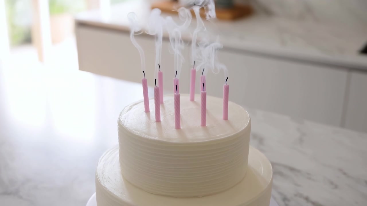 Torta liscia di panna al burro bianca con sottili striature poggia su un piano di lavoro in marmo in una cucina moderna e luminosa, mentre nove candele rosa a spirale si sono appena spente e un sottile fumo sale delicatamente, con torta e candele.