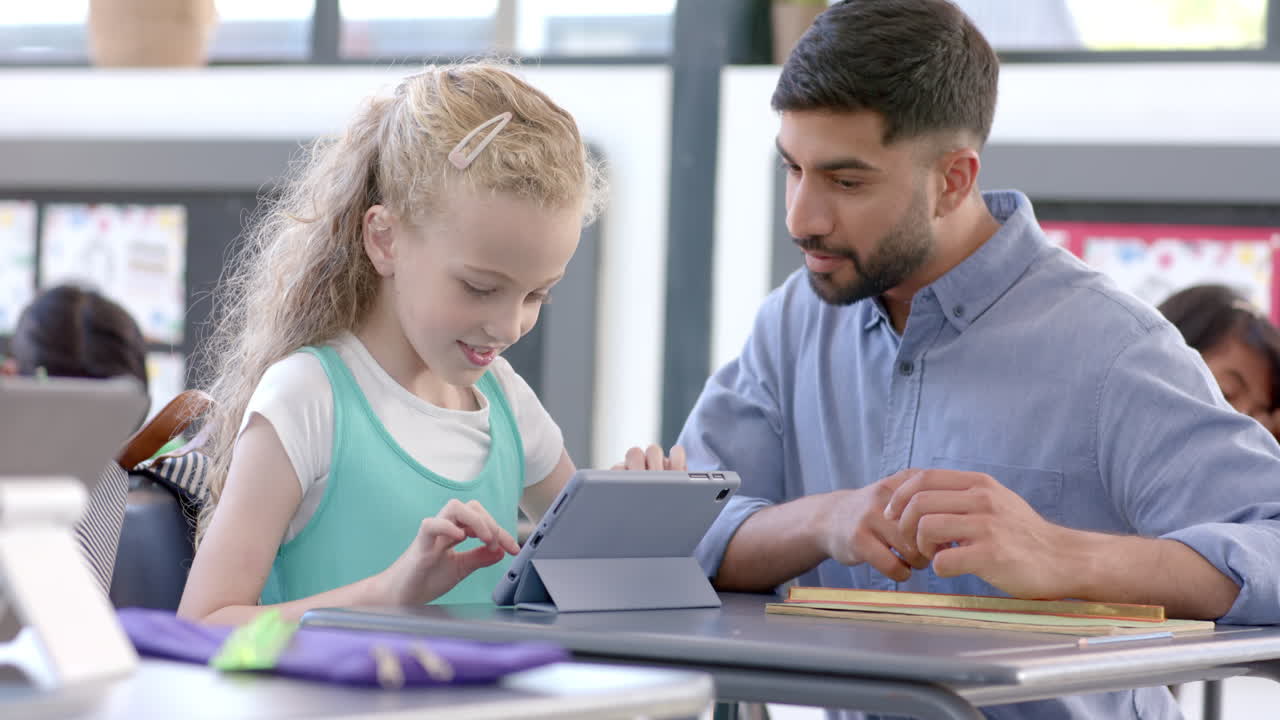 Young Asian man teaches a Caucasian girl at school in a classroom with a tablet