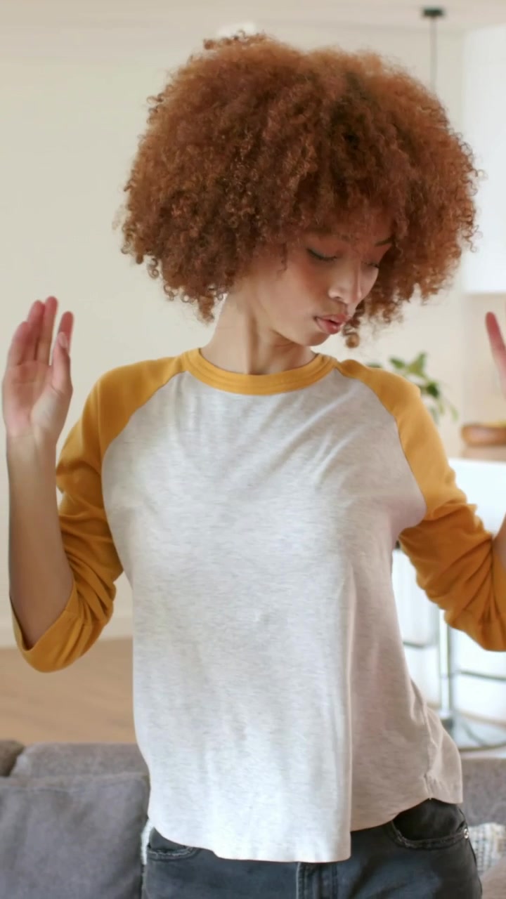 Tanzende Frau in weißem Raglan-Shirt reagiert auf einen Ellbogen außerhalb des Bildes, verschränkt die Arme im Wohnzimmer mit Sofa.