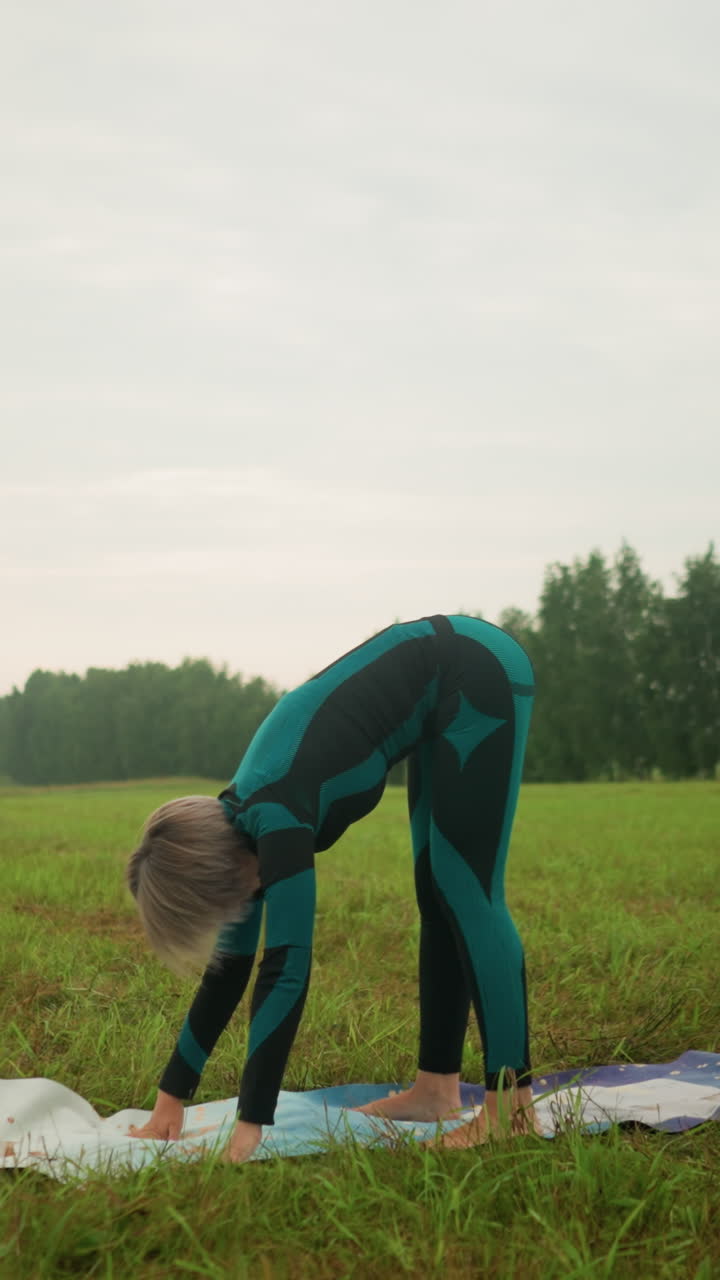 mujer en traje verde y negro realizando la postura del perro mirando hacia abajo, practicando yoga al aire libre en un campo cubierto de hierba bajo un cielo nublado, centrándose en el equilibrio, la flexibilidad y el estiramiento