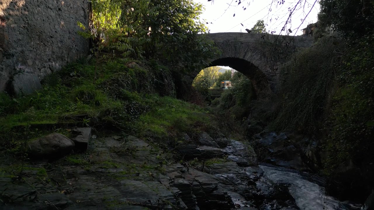 Historic Ponte de Éntoma seen across a shaded stream, with its single stone arch framed by dense vegetation along the Camino de Invierno in Valdeorras, Galicia, Spain