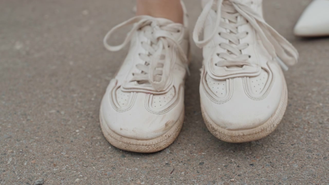 Kvinde binder snørebåndene på en hvid sneaker på ru betongrund, med tæt fokus på hænder og skodetaljer. Handlingen er bevidst, idet kvinden fører snørebåndet gennem snørehullerne for at fastgøre fodtøjet. Blød