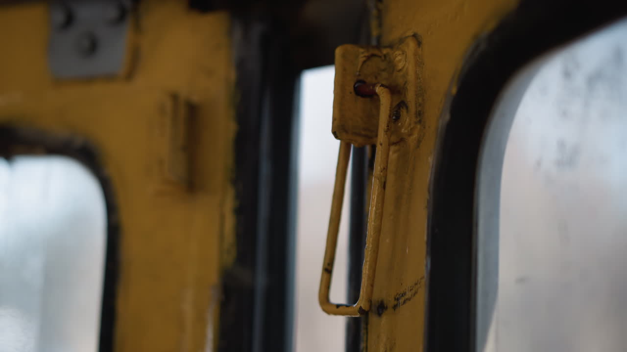 close up view of bus door handle dangling during motion, yellow metal bracket with chipped paint shakes near window, interior transit detail with winter blur outside, urban public transport mood