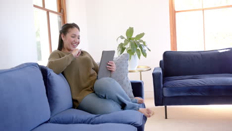 happy biracial woman sitting on sofa, smiling and using tablet in sunny living room