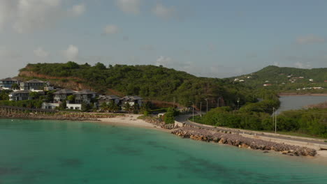 Vista-Aérea-Escénica-Antigua-Y-Las-Bermudas-Costa-Turquesa-Cristalina-Con-Carretera-Asfaltada-Costera-Y-Edificios-Inmobiliarios-De-Lujo-En-Un-Día-Soleado-En-El-Caribe