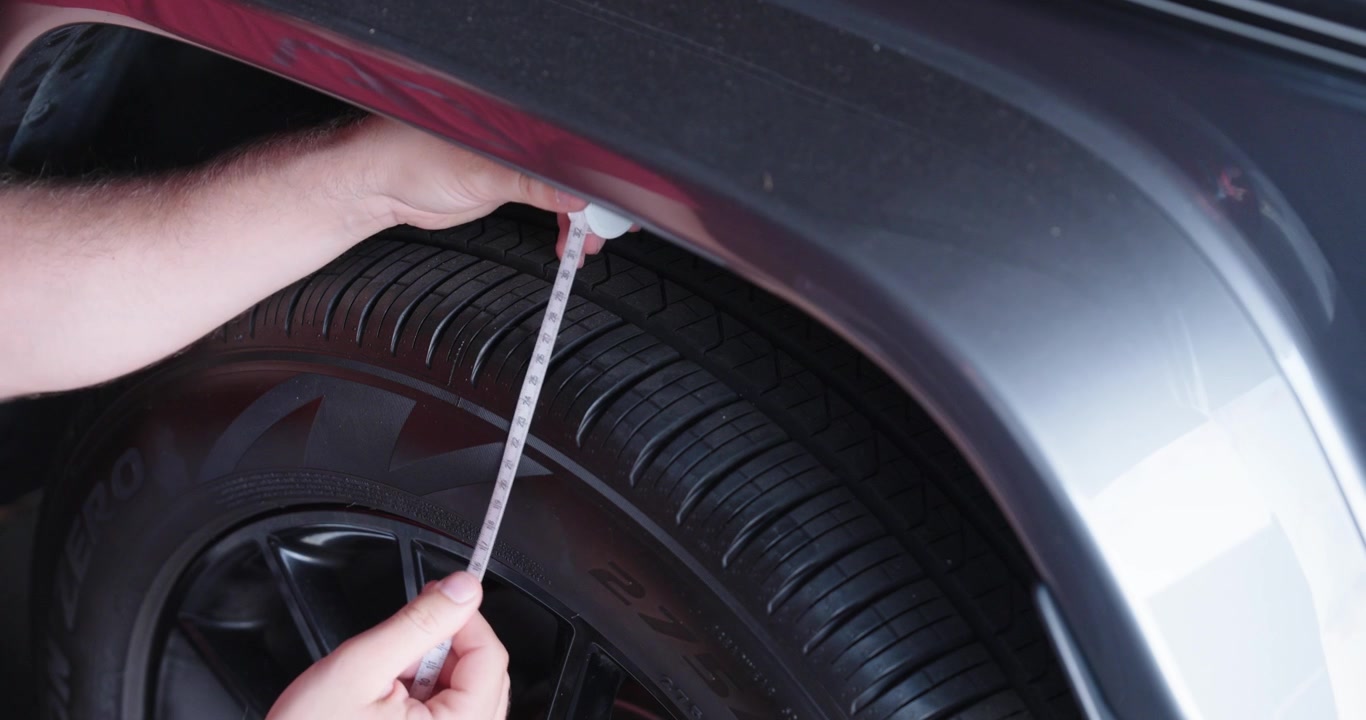 Person measuring tire clearance with a tape measure, checking wheel spacing and vehicle dimensions during inspection.