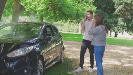 Pareja-Fumando-Coche-Inspeccionando-Los-Daños-Del-Accidente-Y-Llamando-A-Los-Servicios-De-Emergencia