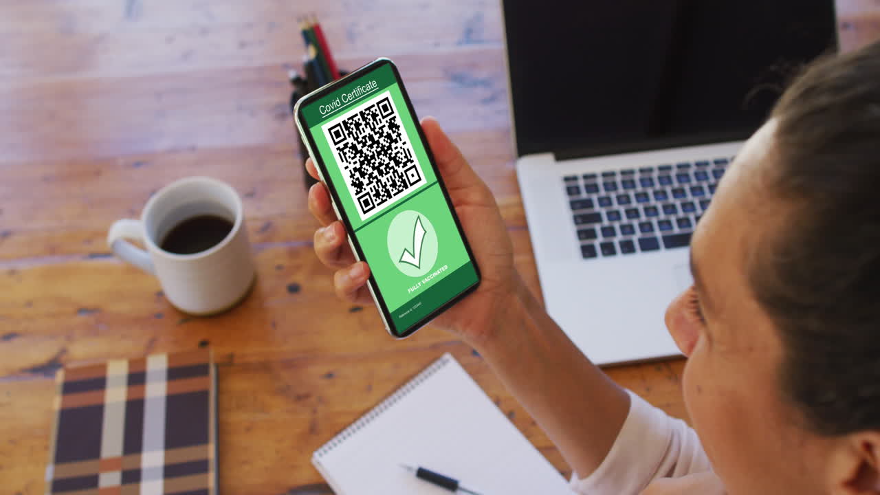 Woman at desk holding smartphone with covid vaccination certificate and qr code on screen