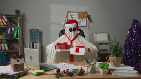 a ghost wearing a santa hat and sunglasses sits at an office desk with christmas decorations, holding gifts