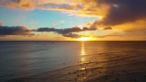 colorful sun setting over group of surfers and boats sailing at dawn on waikiki beach in honolulu hawaii with calm ocean waves - aerial drone shot