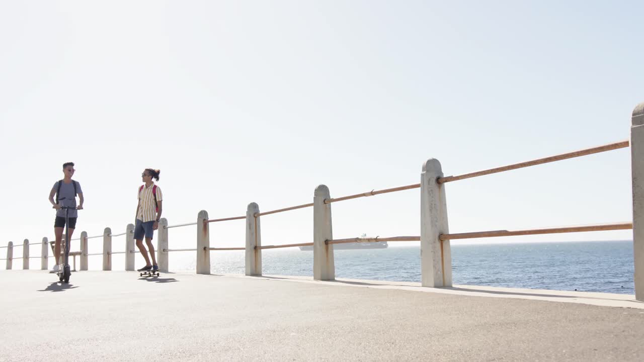 해변의 산책로에서 스쿠터를 사용하는 행복한 다양한 동성애 남성 커플, 슬로우 모션