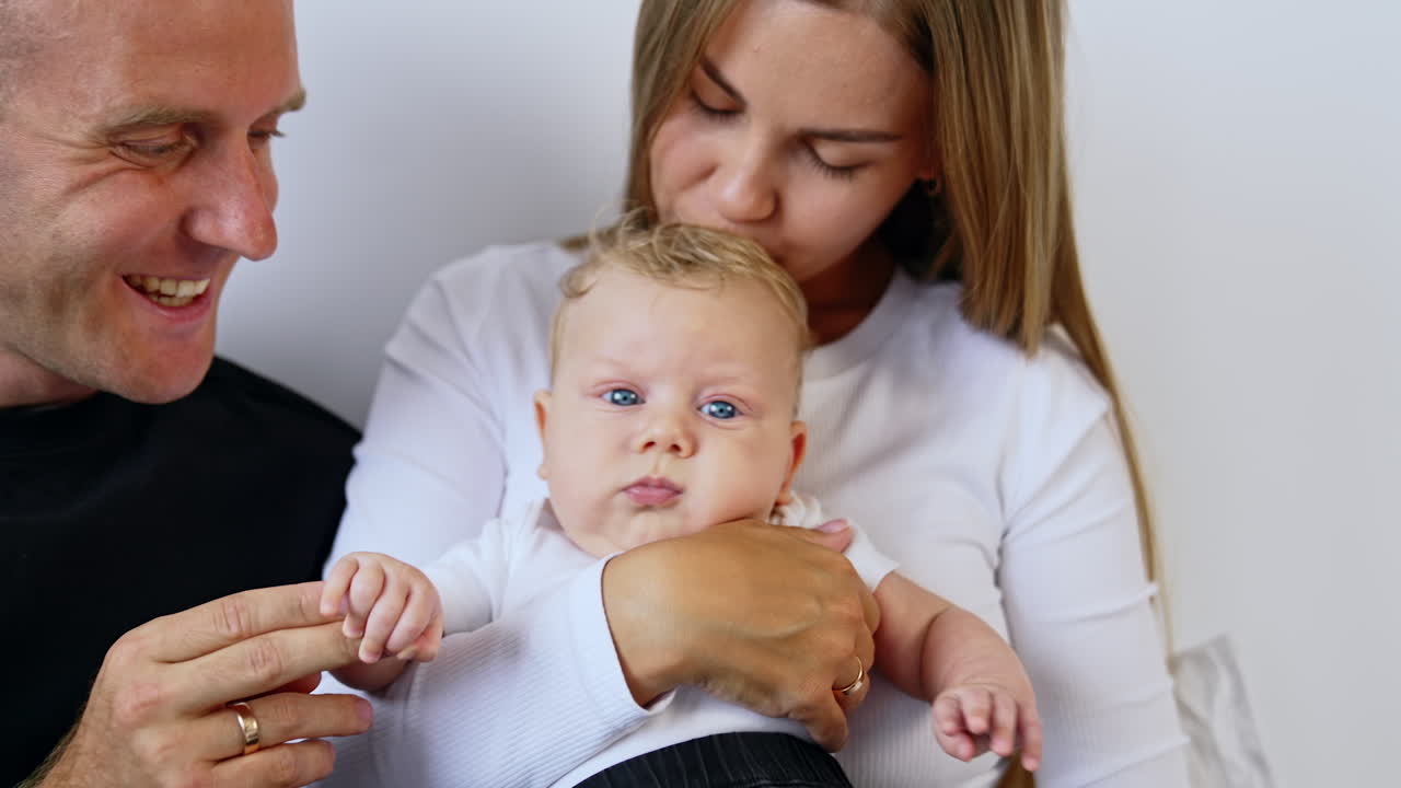 Caucasian woman holding her beautiful blond blue-eyed baby. Mom kisses her infant on the head and dad holds kid's hand.