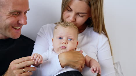 caucasian woman holding her beautiful blond blue-eyed baby. mom kisses her infant on the head and dad holds kid's hand.