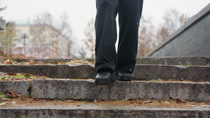 Legs of person walking down stone steps during light snowfall, wearing black jeans and black boots, with colorful autumn leaves scattered along the path